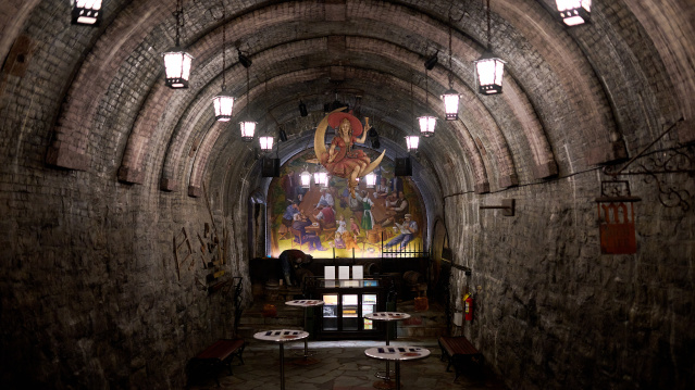 Interior of the historic Miller Caves beneath the Miller Brewery, featuring a long arched brick tunnel with hanging lanterns, stone walls, high-top tasting tables and a colorful brewing-themed mural at the far end.