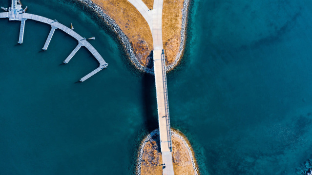 Aerial view of a pedestrian bridge stretching over deep blue water, connecting two small land areas with golden-brown grass. To the left, empty docks extend into the water, creating geometric shapes against the shoreline.