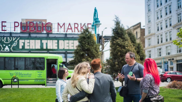 A small group gathers in a downtown park during a guided tour, listening as a guide speaks. A green Milwaukee Food & City Tours bus and the iconic Milwaukee Public Market sign are visible in the background, highlighting the city's vibrant Third Ward district.