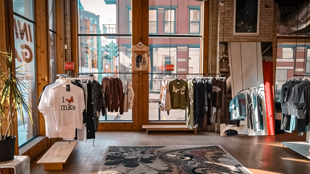 Interior of Unfinished Legacy clothing store in Milwaukee, featuring streetwear displayed on minimalist racks, large sunlit windows, exposed brick walls, and locally inspired graphic apparel including Milwaukee-themed T-shirts.