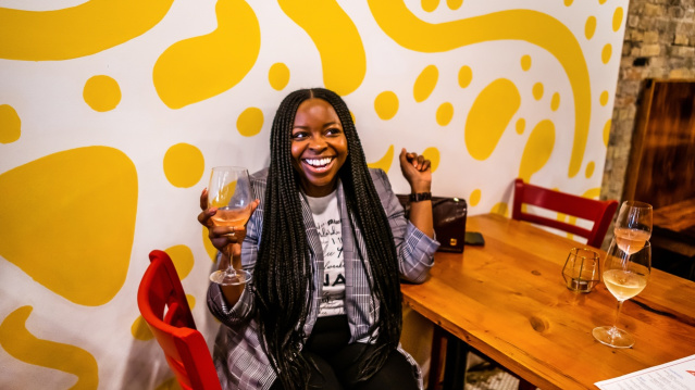 a woman sitting in front of a yellow and white patterned wall laughing and smiling while holding a glass of white wine