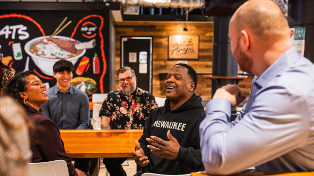 LeRoy Butler is wearing a black Milwaukee hoodie speaks animatedly while seated at a table inside 3rd Street Market Hall, with several people listening and smiling around him