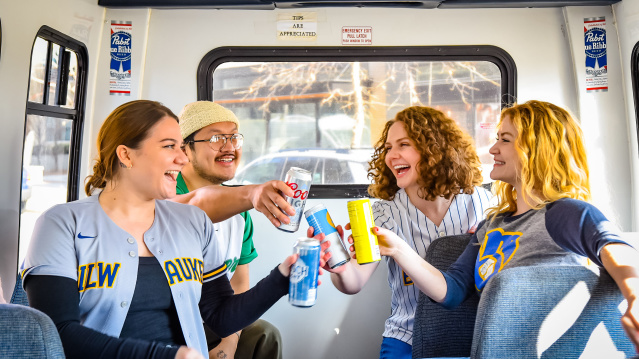 Four friends sitting inside a shuttle bus, smiling and clinking their drinks together. Two are wearing Milwaukee sports jerseys, creating a festive and celebratory atmosphere.