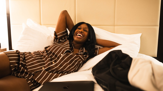 A person lying on a hotel bed, smiling and laughing with their arms resting behind their head. A closed laptop sits on the bed nearby, and a jacket is draped beside them. The room has soft lighting and a padded headboard, creating a relaxed, comfortable atmosphere.