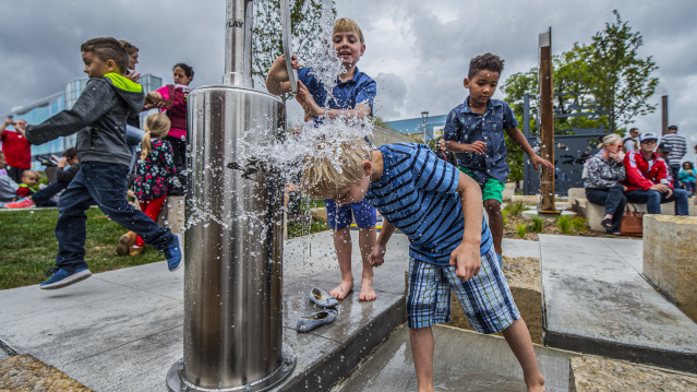 Children play joyfully at an outdoor water pump, one boy splashes under the water, others giggle nearby. Cloudy sky and spectators in the background.
