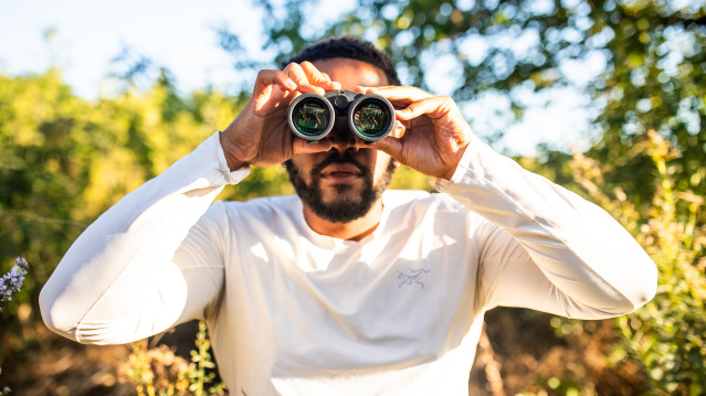 A man in a white long-sleeved shirt holds binoculars up to his eyes while standing outdoors in a sunny, tree-filled setting with green foliage and dappled sunlight in the background.