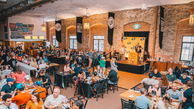 An open banquet hall filled with people eating food and drinking beer while a performer plays the accordion in the background.