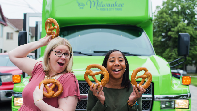 two friends standing in front of green tour bus showing off soft pretzels