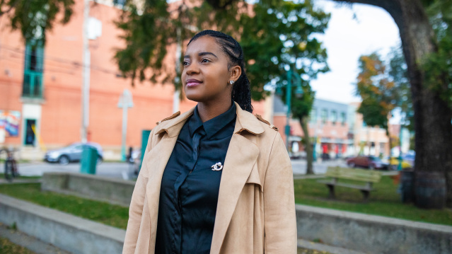 Ashley Jordan stands outdoors in an urban setting, wearing a tan trench coat over a dark shirt, with braided hair and a slight smile, with brick buildings and trees visible in the background