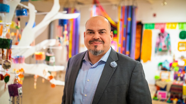Jacobo Lovo of Latino Arts stands smiling in a colorful community space, wearing a gray blazer and light blue shirt with a decorative pin
