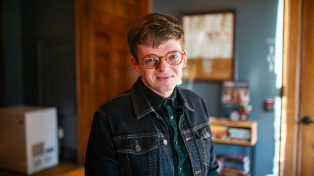 Ryan Castelaz with short brown hair and round glasses smiles warmly at the camera. They're wearing a dark denim jacket over a dark green plaid shirt, standing in what appears to be a cozy indoor space with warm wood tones, bookshelves, and soft lighting in the background.