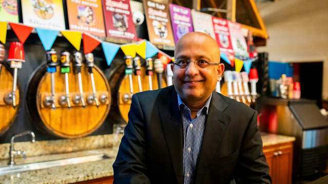 Sharad Chadha, CEO of Sprecher Brewing Company, sits smiling in a dark suit and blue checkered shirt at the brewery's taproom bar. Behind him are multiple beer taps, colorful triangular bunting, and shelves displaying various Sprecher product bottles and signage.