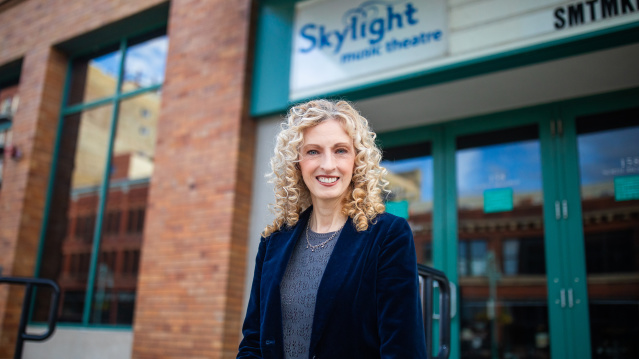 Susan Varela, executive director of Skylight Music Theatre, smiles while standing in front of the theater's brick building with the Skylight music theatre sign visible in the background