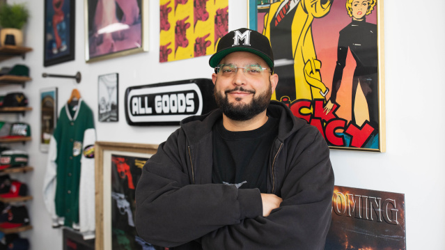 headshot of a latino man with a beard smirking in front of a colorful gallery wall with a ball cap on and his arms crossed
