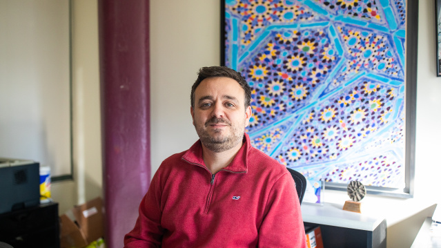 Milwaukee Rep Director Chad Bauman wears a red shirt and sits with hands clasped in his office with geometric artwork in the background.