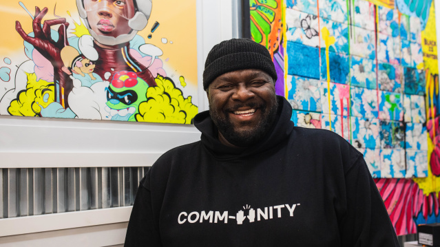 a portrait of a black man laughing in front of a gallery wall of colorful art with a black beanie and black hoodie that reads "COMMUNITY" on it in white lettering