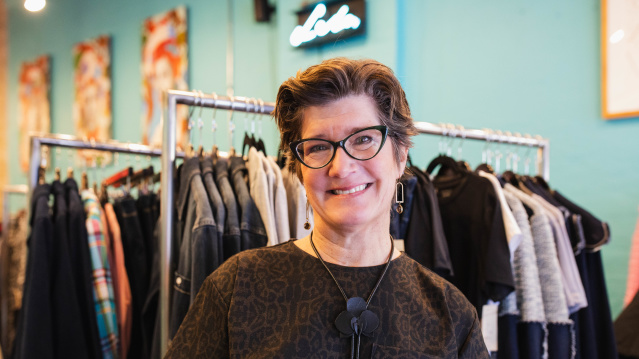 a portrait of a white woman with cat eye glasses smiling in front of a rack of clothes