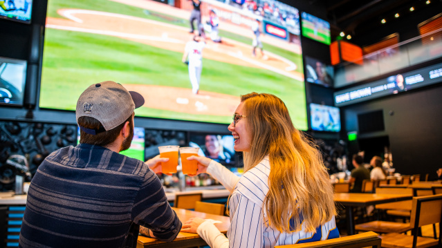 A man and woman clinking glasses of beer while watching baseball