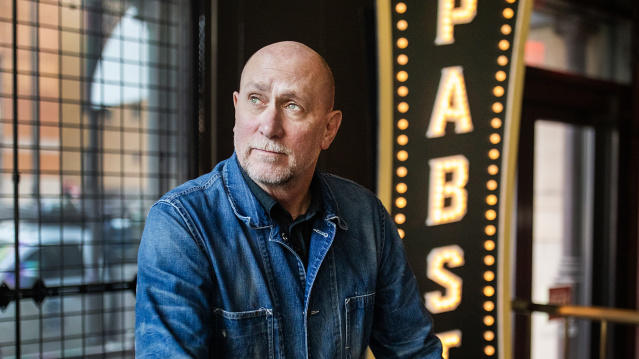 Gary Witt sits wearing a denim shirt in front of an illuminated Pabst sign with vintage-style light bulbs, with large windows visible in the background