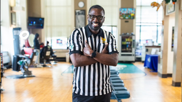 Gaulien 'Gee' Smith, a Black man wearing glasses and a black and white striped referee shirt, stands with his arms crossed and a big smile in what appears to be a gym or recreation center. The polished wooden floor and exercise equipment are visible in the background, along with large windows and colorful wall decorations.