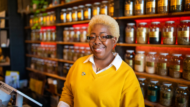 Angela Mallett is smiling, standing behind the counter of HoneyBee Sage. She's wearing a bright yellow sweater over a white collared shirt, along with gold jewelry including earrings and a necklace. Behind her are wooden shelves lined with numerous glass jars containing various spices, herbs, or other ingredients with labels.