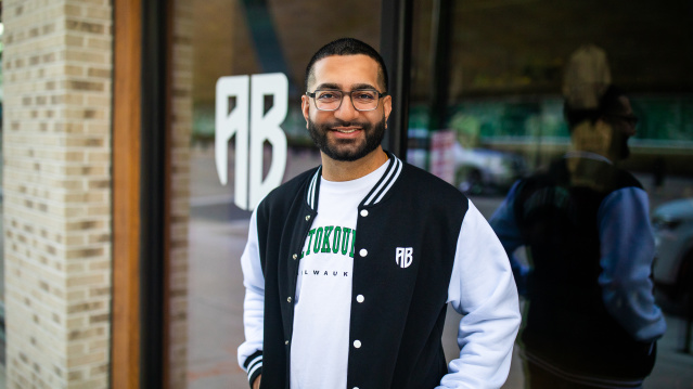 Nimesh "Nemo" Ruparel stands smiling outside a storefront wearing a black and white varsity-style jacket with "MILWAUKEE" text in green letters and "AB" logo. He has glasses, a beard, and is positioned in front of glass windows with "AB" lettering visible in the background. The setting appears to be an urban commercial area with brick architecture.