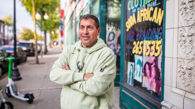 Peter Feigin in a light green hoodie with a deer logo stands with his arms crossed, smiling at the camera on a city sidewalk. Behind him is a colorful storefront window with "CLOTHES" and other text visible, along with decorative stone architecture. Trees and parked cars line the urban street scene.