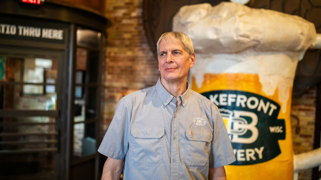 Russ Klisch wears a light blue button-down shirt at Lakefront Brewery. Behind him are exposed brick walls, industrial-style lighting, and a large format frothing beer mug.