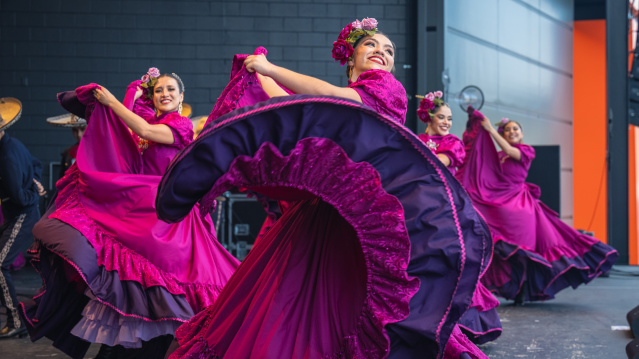 A group of female dancers performs a traditional Mexican folklórico dance on stage. They wear vibrant magenta dresses with purple ruffles and floral headpieces. The lead dancer in the foreground smiles brightly while gracefully twirling her flowing skirt. The dancers behind her mirror the movement, creating a colorful, energetic scene against a modern stage backdrop.