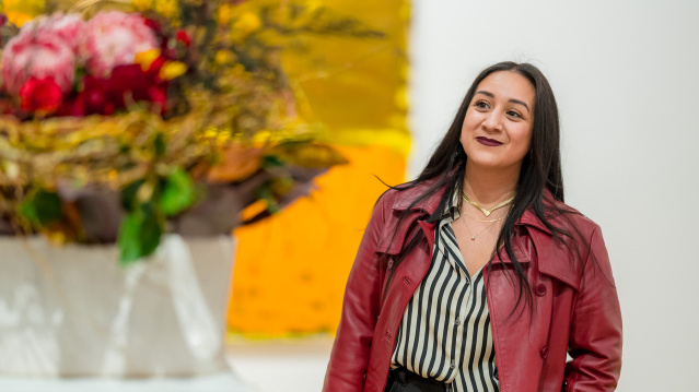 Woman in a red leather jacket and striped blouse smiling while looking at a large floral arrangement, with bright yellow artwork in the background.