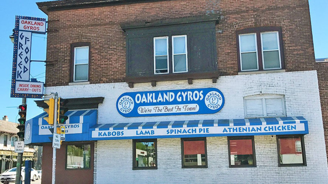 Front view of Oakland Gyros, a two-story brick building with a white-painted lower level and blue signage reading “Oakland Gyros – Best Gyros in Town.” The storefront features blue awnings advertising kabobs, lamb, spinach pie, and Athenian chicken