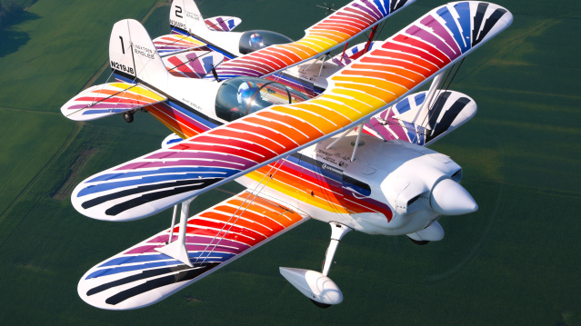 Two brightly colored biplanes fly in close formation above green water, showcasing coordinated aerobatic precision. Their vibrant red, orange, blue, and white striped wings create a striking visual display against the calm background below.