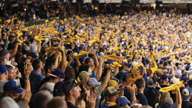 A packed stadium crowd cheers and waves yellow rally towels during a baseball game, filling the stands with motion and team colors.