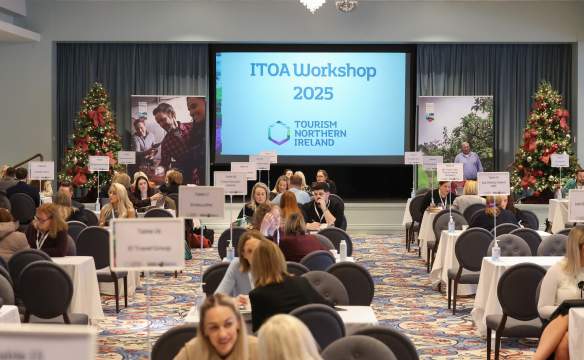Delegates take part in one-to-one business meetings at the ITOA Workshop 2025, seated at numbered tables in a conference venue. A large screen displaying “ITOA Workshop 2025” and Tourism Northern Ireland branding is visible at the front of the room, with exhibition banners and decorated Christmas trees on either side.