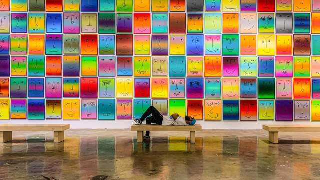 A man laying on a bench in front of the Happy Exhibit at NSU Art Museum