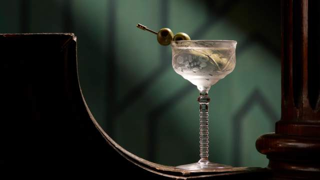 A classic martini in an etched coupe glass sits on the curved edge of a dark wooden surface, garnished with two green olives on a cocktail pick. Soft, moody lighting highlights the clear drink and glass stem against a deep green, art deco–style background.