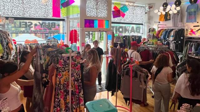 A bright, colorful vintage and clothing shop filled with racks of patterned dresses, shirts, and accessories. Several shoppers browse the tightly packed displays, and natural light comes in through large front windows decorated with bold, geometric designs. The space has a lively, eclectic atmosphere with playful décor and a mix of retro and modern styles.