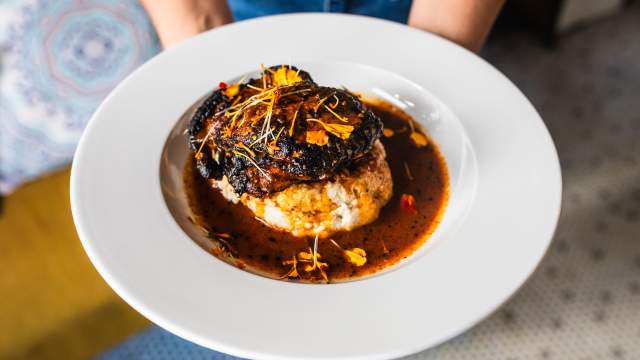A beautifully plated dish featuring a charred, glazed piece of meat resting atop a fluffy biscuit, surrounded by a rich brown sauce and garnished with delicate edible flower petals.