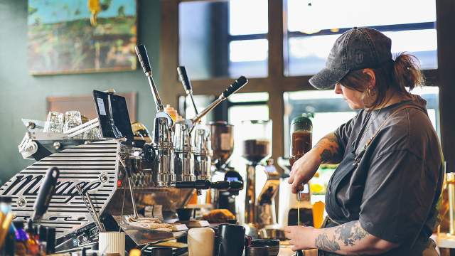 Barista preparing a drink at a coffee machine inside a café, surrounded by coffee-making equipment and supplies.