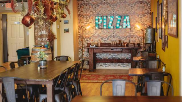 Interior of Flour Girl & Flame with wooden tables, metal chairs, hanging copper pots and a decorative wall with a bright “PIZZA” sign.