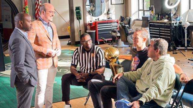 Five men gather in a barbershop setting, with one man in a referee shirt holding a golden NBA championship trophy while others look on. The scene includes professional barber equipment, mirrors, and appears to be during a casual interview.