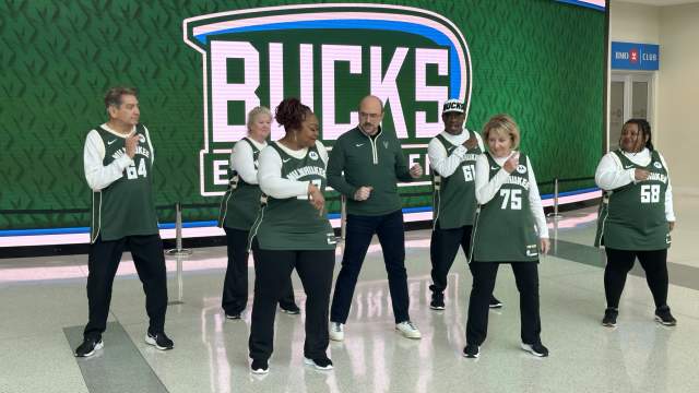 A group of Milwaukee Bucks Grand Dancers poses in the concourse area of Fiserv Forum in front of a large green wall featuring the Bucks logo. The seven dancers, wearing matching green and white Milwaukee Bucks jerseys with numbers like 34, 61, and 75, stand alongside what appears to be a team official or coach in a green quarter-zip pullover. The polished concrete floor reflects the overhead lighting, and BMO Club signage is visible in the background, creating a professional arena atmosphere.