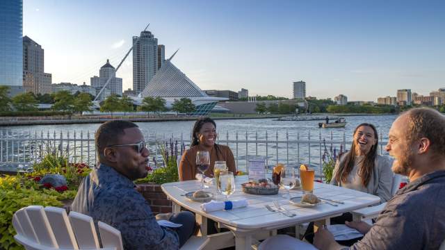 Four friends dining outdoors at Harbor House in Milwaukee, enjoying drinks and seafood by Lake Michigan with the Milwaukee Art Museum and city skyline in the background.