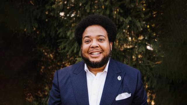 Jonathan Newby smiles at the camera while wearing a navy suit and white shirt. He is standing in front of greenery.