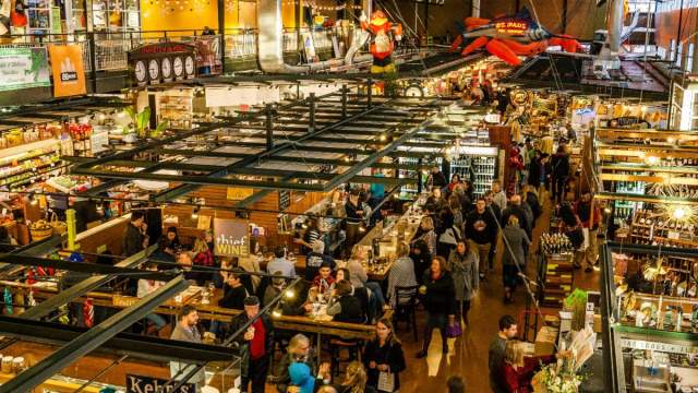 A bustling indoor public market filled with shoppers walking through narrow aisles lined with food vendors, produce stands, and specialty shops. People sit at crowded counters and tables enjoying food and drinks. Overhead, colorful decorations and large hanging sculptures add to the lively, energetic atmosphere.