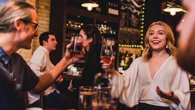 A lively bar scene where a smiling woman holding a glass of wine gestures warmly while talking to others. People around her raise drinks in a toast, and small groups sit and chat in the cozy, dimly lit space filled with bottles, chalkboard menus, and warm ambient lighting.