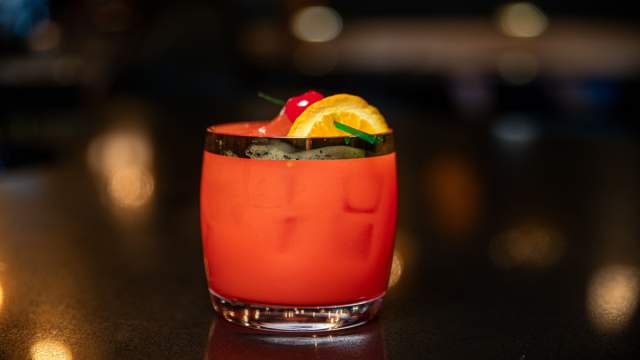 A vibrant red Rum Punch cocktail served in a rocks glass, garnished with an orange slice and maraschino cherry, set on a dark bar surface with warm, blurred lights creating bokeh in the background.