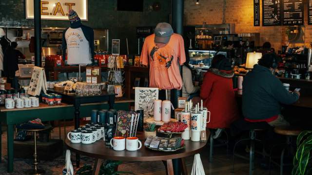 Interior of a trendy café with a round display table of merchandise like mugs, shirts, and tote bags in the foreground, and people seated at the counter enjoying drinks in the background.