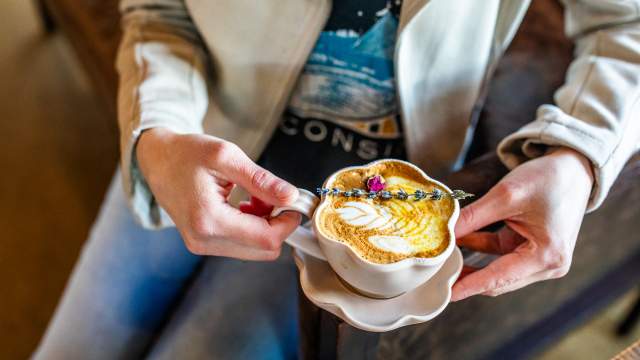 A person holds a white coffee cup and saucer with both hands, presenting a latte with intricate leaf-pattern latte art in golden-brown foam. The coffee is garnished with a small sprig of lavender and a pink flower petal. The person is wearing a white long-sleeved jacket, and the background shows a warm, blurred cafe setting.