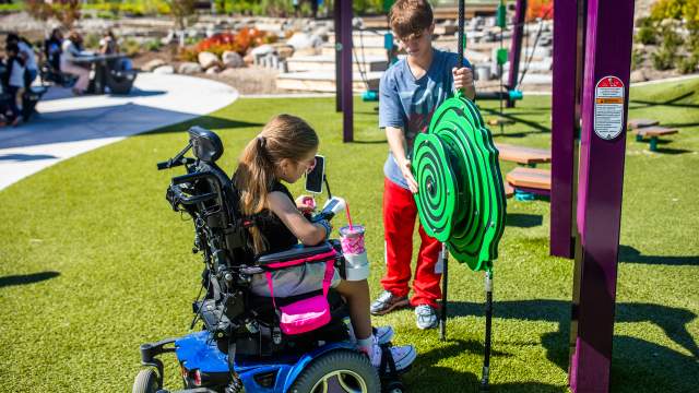 A child using a power wheelchair plays with an interactive spinner at an accessible playground while another child stands beside them, helping turn the green wheel, with landscaped paths and play structures in the background.